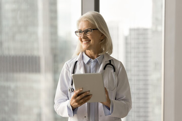 Cheerful mature practitioner woman in glasses and white coat using tablet computer for job...