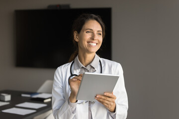 Positive adult doctor woman using tablet computer in office conference room, looking away with...