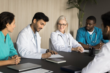 Diverse team of medical professionals making business decision on meeting with colleagues, partners. Confident young Indian chief doctor signing agreement, hiring new specialist