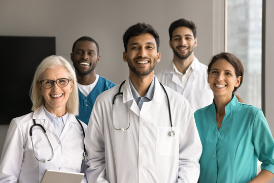 Happy young Indian medical boss and diverse team of doctors, practitioners, surgeons in uniforms looking at camera with toothy smiles, posing for portrait. Multiethnic small clinic staff shot