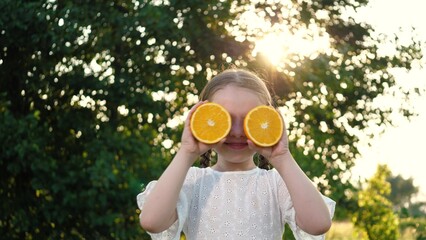 Joyful little girl holds orange halves by face in summer park exotic fruits