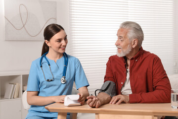 Fototapeta premium Young healthcare worker measuring senior man's blood pressure at wooden table indoors