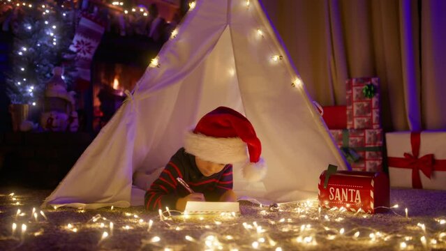 Funny kid in large red Santa hat comfortably lying in tent with magic light garland. Cute little boy writing wishes in letter to Santa Claus in fairy tale atmosphere in living room with fireplace 4K