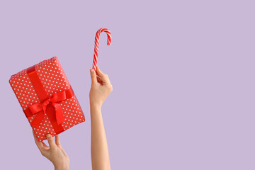 Female hands holding Christmas gift and candy cane on lilac background