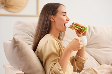 Beautiful young woman with cup of coffee eating tasty sandwich while resting on sofa in living room