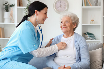 Female nurse hearing senior woman with stethoscope at home