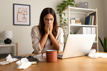 Sick woman working from home office. Caucasian female blowing nose with tissue while working with laptop.
