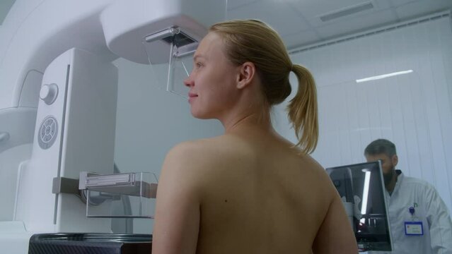 Happy Caucasian woman stands topless in clinic radiology room. Female patient undergoing mammography scanning examination in modern hospital. Radiologist adjusts mammogram machine using computer.