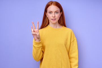 beautiful happy cheerful redhead woman in yellow stylish sweater demonstrating letter N sign language symbol for deaf human with blue background. isolated close up portrait
