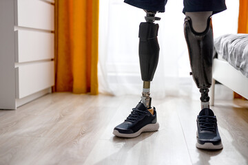 handicapped young unrecognizable man with leg prosthesis in sneakers standing indoors at home,...