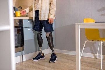 cropped handicapped or disabled person with disability standing on bionic smart prosthesis in kitchen, making coffee or tea for himself at home. indoors. in the morning. disability lifestyle