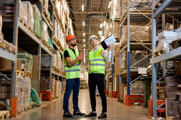 warehouse managers in work wear organizing distribution in warehouse storage area. Caucasian men in green uniform discuss product delivery, having talk. in retail Warehouse full of Shelves with Goods
