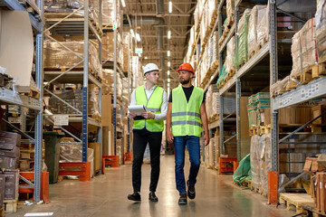 two men engineers in safety helmets and green uniforms walking among shelves with goods in warehouse talking, checking goods. logistic and business export, distribution and inventory concept