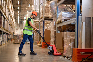 male staff worker in warehouse uses hand pallet stacker to transport goods, alone, dressed in working clothes and safety hard hat.Skilled warehouse employee pushing manual pallet jack - obrazy, fototapety, plakaty
