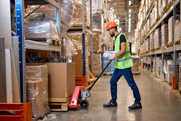 caucasian worker in a warehouse uses hand pallet stacker to transport pallets good cargo, hardworking confident male staff in green uniform vest at work place. Hand pallet truck jack in warehouse.