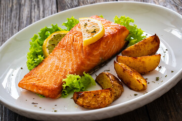 Seared salmon steak with baked potatoes and fresh vegetable salad served on wooden table
