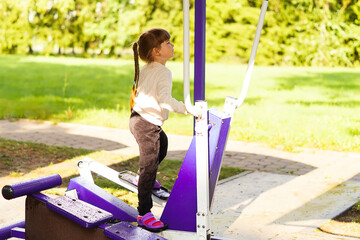 A funny five-year-old girl is doing street fitness on a simulator. A little beautiful girl does exercises on a street sports ground. Healthy childhood concept. Sports and children. City sports ground