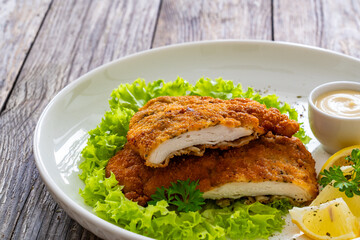 Crispy breaded seared chicken cutlet with lemon slices and fresh vegetables on wooden table
