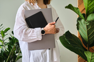 A woman holds a laptop and graphics tablet against a background of greenery.