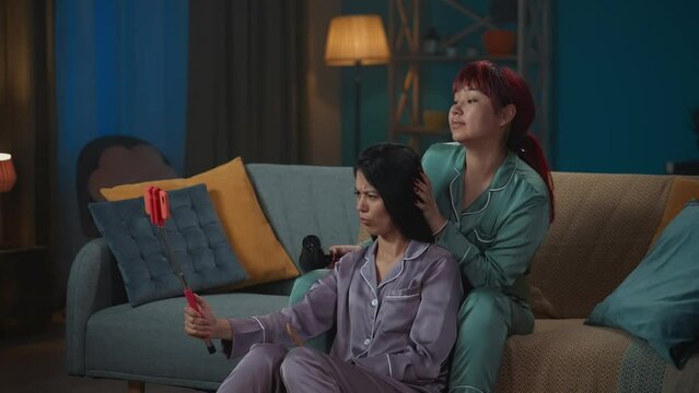 Medium-full video capturing two young women wearing pajamas sitting on the couch, taking selfies using a selfie stick as one of them is doing the other one's hair.