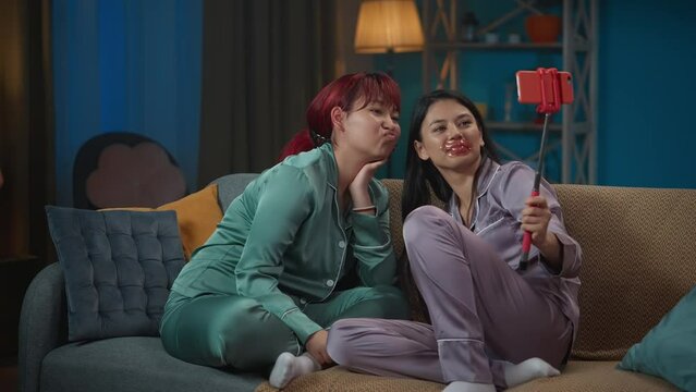 Medium-full video capturing two young women wearing pajamas sitting on the couch and doing their evening skincare routine, taking photos using a selfie stick.