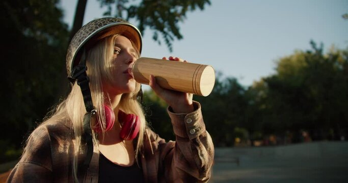 Close-up shot of a blonde girl in a leopard print helmet in a plaid shirt and black top drinking water from a sports bottle while relaxing in a skatepark in the summer