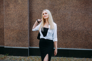 Fototapeta na wymiar A beautiful smiling slender woman with long blond hair, in a black long dress and a white shirt, stands on a city street, against the backdrop of a granite building wall.