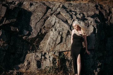 A slender, positive, sexy aged woman with long blond hair in a black long stylish dress stands near the picturesque rocks on a bright sunny summer day.