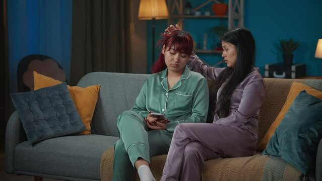 Medium-full video capturing two young women in pajamas sitting on the couch together. One of them is sad about something in her smartphone, her friend, sister thies to cheer her up.
