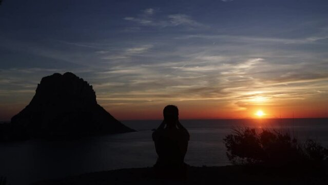 A man sitting on the ground enjoying the sunset and taking photos with his mobile phone.