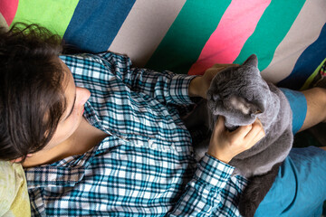 A teenager is lying on the couch at home and stroking a gray Scottish cat.