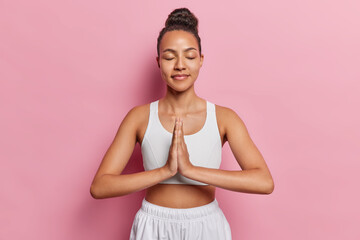 Photo of sporty Latin woman with dark hair gathered in bun meditates with her eyes closed keeps palms in praying gesture maintains healthy lifestyle practices yoga isolated over pink background