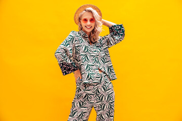 Young beautiful smiling female in trendy summer hippie costume clothes. Carefree woman posing near yellow wall in studio. Positive blond model having fun. Cheerful and happy. In hat and sunglasses