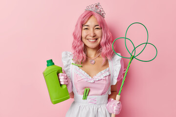 Horizontal shot of cheerful Asian maid wears crown and white dress holds bottle of cleaning detergent and carpet beater busy with cleaning isolated over pink background. Housekeeping concept