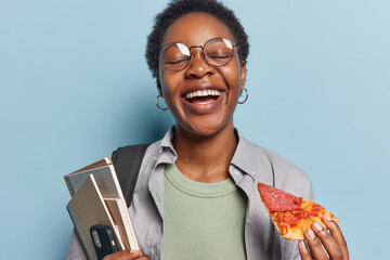Photo of overjoyed clever dark skinned female teenager laughs happily eats tasty pizza and holds textbooks with smartphone cannot stop laughing at something funny poses against blue background