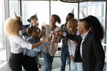 Excited employees celebrating African American businesswoman colleague birthday in office, clinking glasses with champagne and cheering, presenting gift, business people involved in party