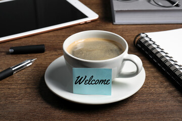 Sticky note with word Welcome attached to cup of coffee on wooden office desk