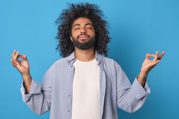 Young relaxed bearded Arabian man takes yoga pose and meditates to clear head of negative thoughts and plunge into zen trance dressed in casual clothes stands on plain blue background.