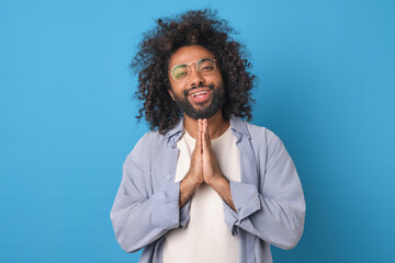 Young kind positive Arabian man smiles and makes prayer gesture demonstrating readiness to forgive offender committed unpleasant act dressed in casual style stands on blue studio background. - obrazy, fototapety, plakaty