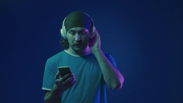 A man in white wireless headphones flips through a playlist on his smartphone, turns on the music and dances. A man in a studio on a blue background in pink and green neon light.