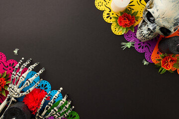 Experience mexican heritage on Dia de los Muertos. Top view shot of skull, skeleton hands, vibrant flowers, candles, paper garland, scary decor on black background with ad place