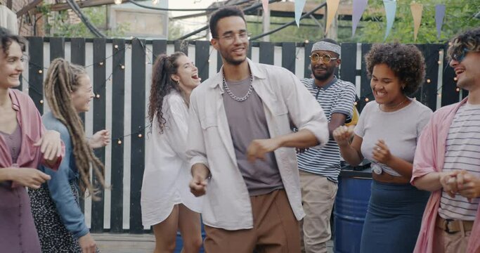 Slow motion of excited young men and women dancing outdoors in street bar listening to DJ music and laughing at party. Youth culture and entertainment concept.