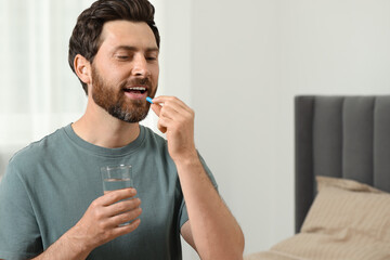 Handsome man with glass of water taking pill at home, space for text - obrazy, fototapety, plakaty