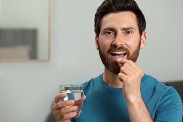 Handsome man with glass of water taking pill indoors, space for text