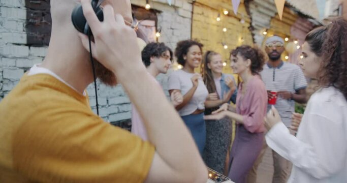 Young man DJ working at modern party while diverse group of people dancing having fun in open air cafe. Entertainment and culture concept.