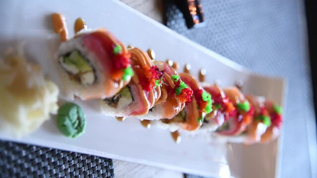 Close-up side view of sushi roll set with salmon and tuna on white plate standing on wooden table in restaurant. Real time handheld vertical video. Japanese food theme.