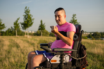 Man wearing pink t-shirt in wheelchair holds smartphone against countryside view. Male with hands...