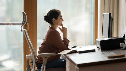 Side view thoughtful Indian businesswoman looking at computer screen, touching chin, sitting at...