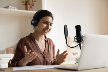 Smiling Indian woman in wireless headphones talking speaking, using professional microphone and laptop, young female engaged in virtual event, successful businesswoman blogger recording podcast