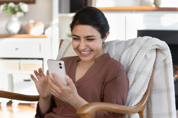 Close up excited Indian woman using smartphone, sitting in cozy chair at home, happy young female...